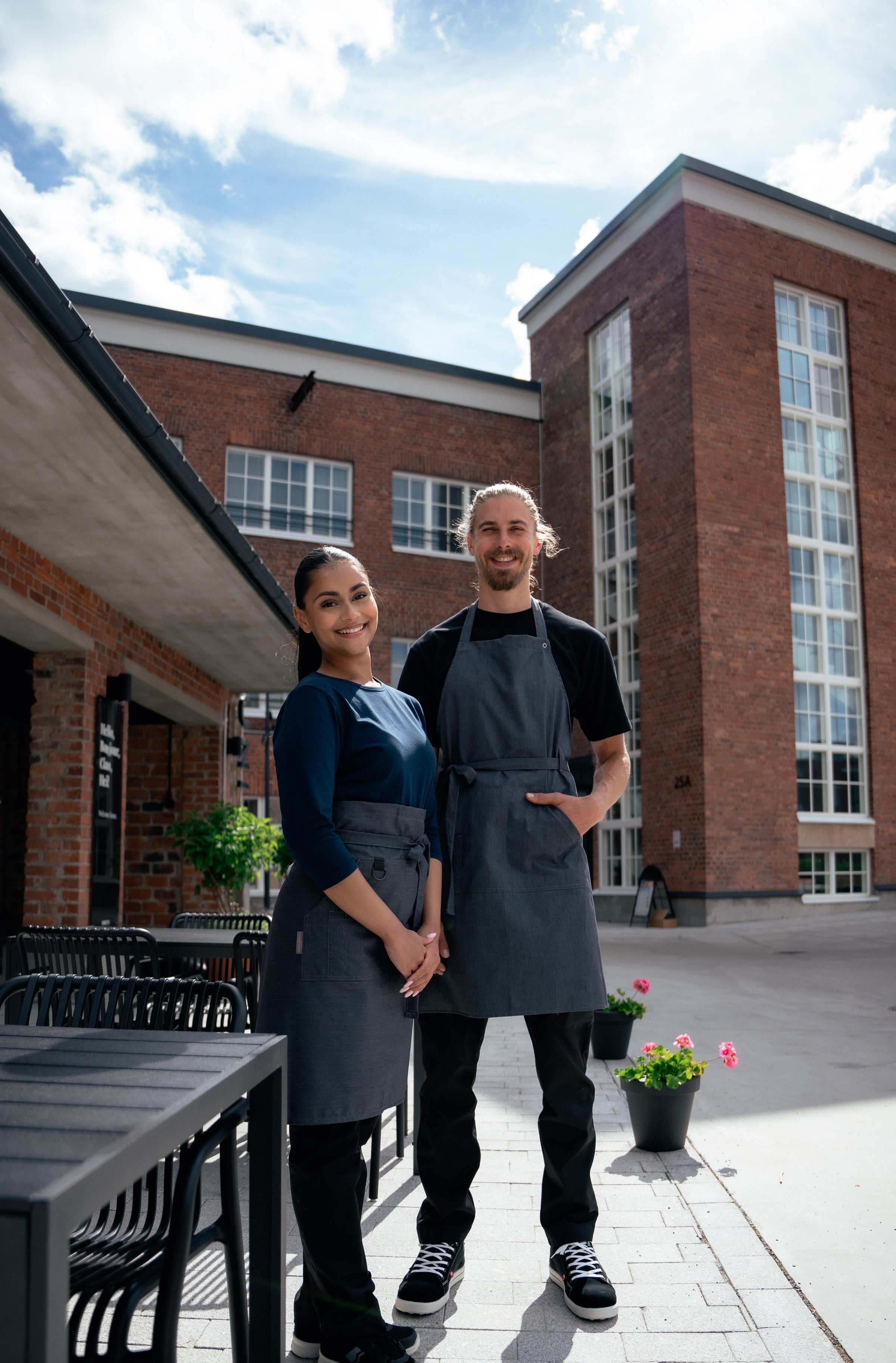 Kuvassa nainen ja mies istuvat ulkona tasolla. Molemmilla on Image Wearin suunnittelemat ja valmistamat mustat joustavat reisitaskuhousut ja työliivi sekä valkoinen T-paita. Jalassa heillä on Sievin Planar turvakengät.