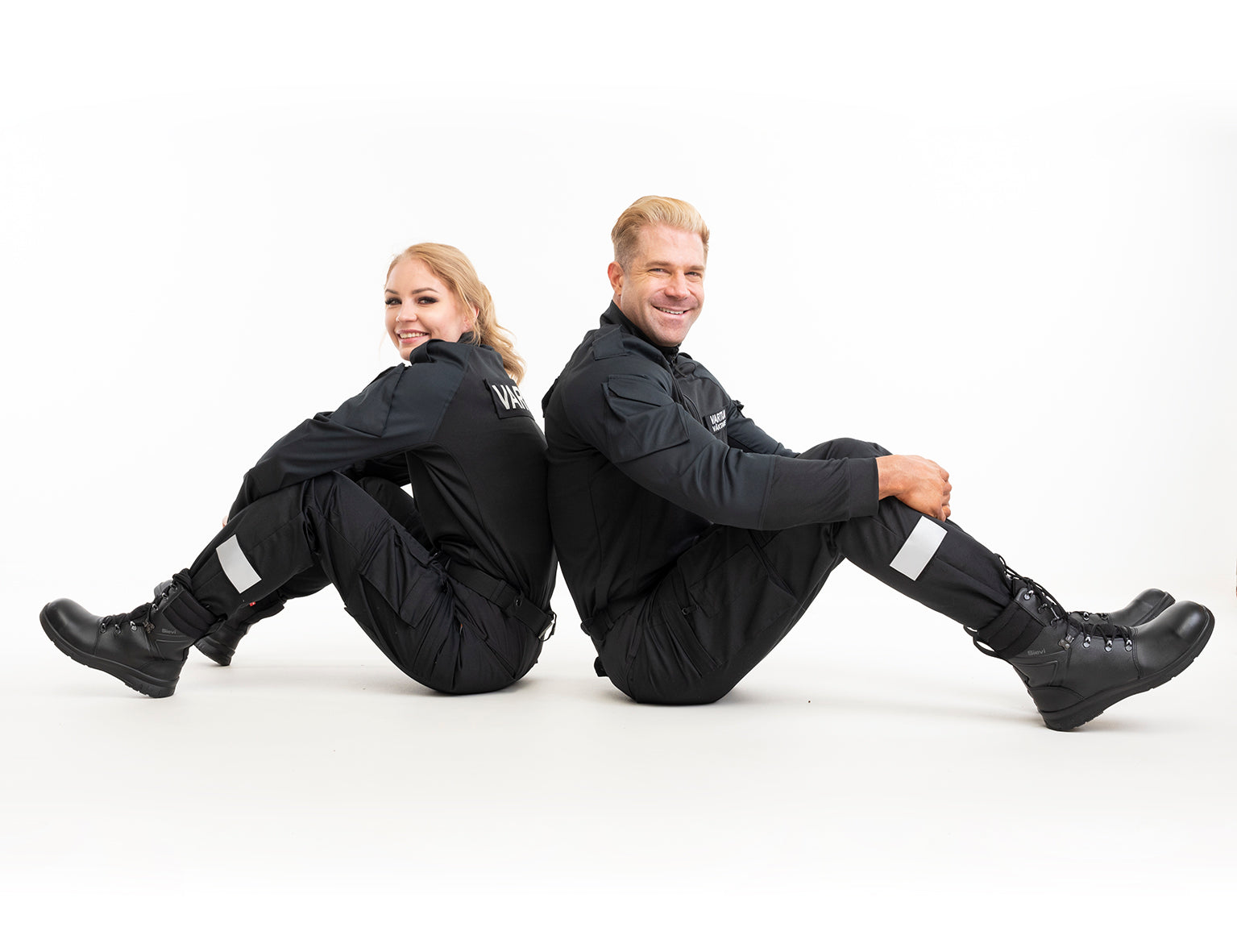 Kuvassa nais- ja miesvartija istuvat lattialla. Molemmilla on päällä Image Wearin malliston Vartija kenttähaalari sekä Sievin varrelliset turvajalkineet..