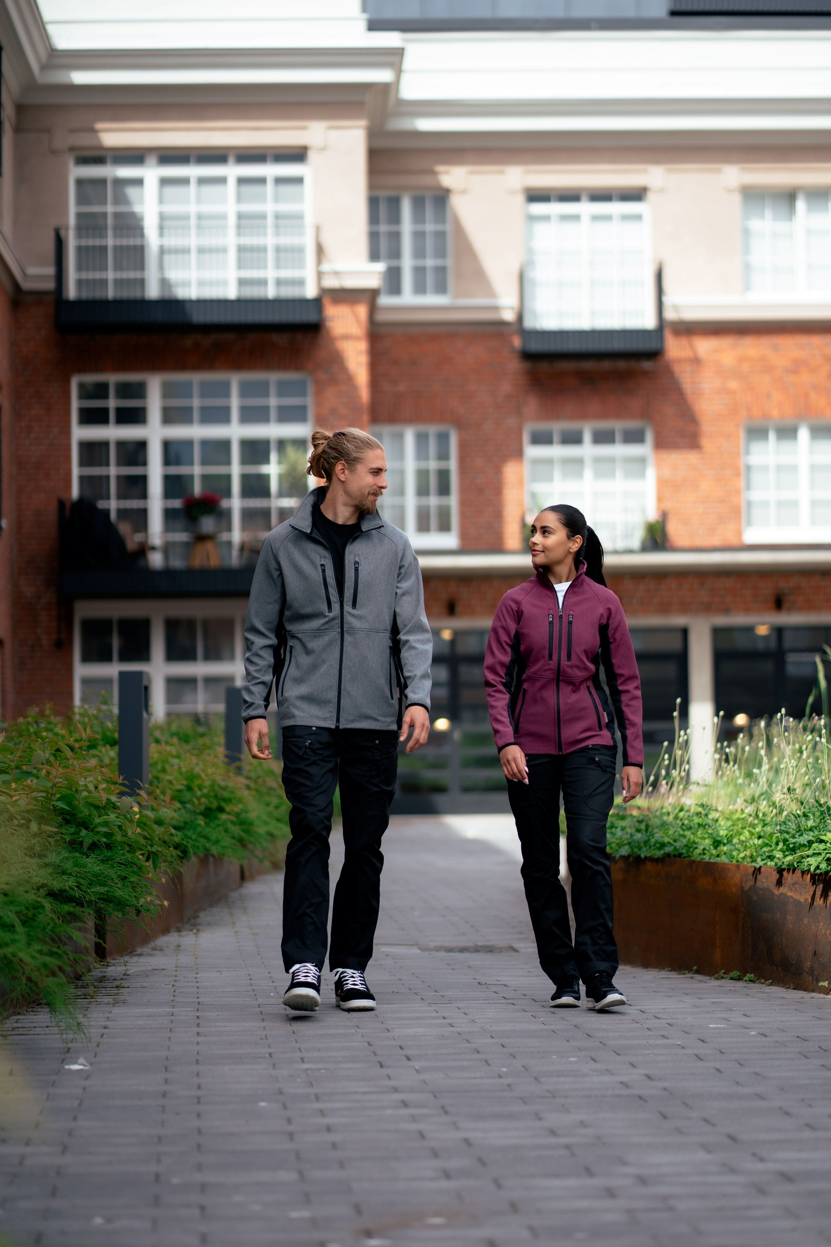 Kuvassa nainen ja mies kävelevät ulkona. Molemmilla on päällä softshell takit Image Wearin valikoimasta ja mustat joustavat työfarkut sekä Sievin työkengät. 