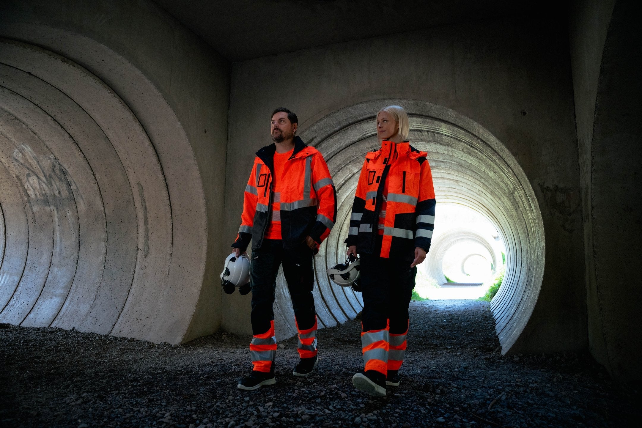 Kuvassa mies ja nainen seisovat betonitunnelissa päällään oranssi varoitusvaatteet, Sievin turvakengät ja Petzlin kypärät Image Wearin mallistosta. 
