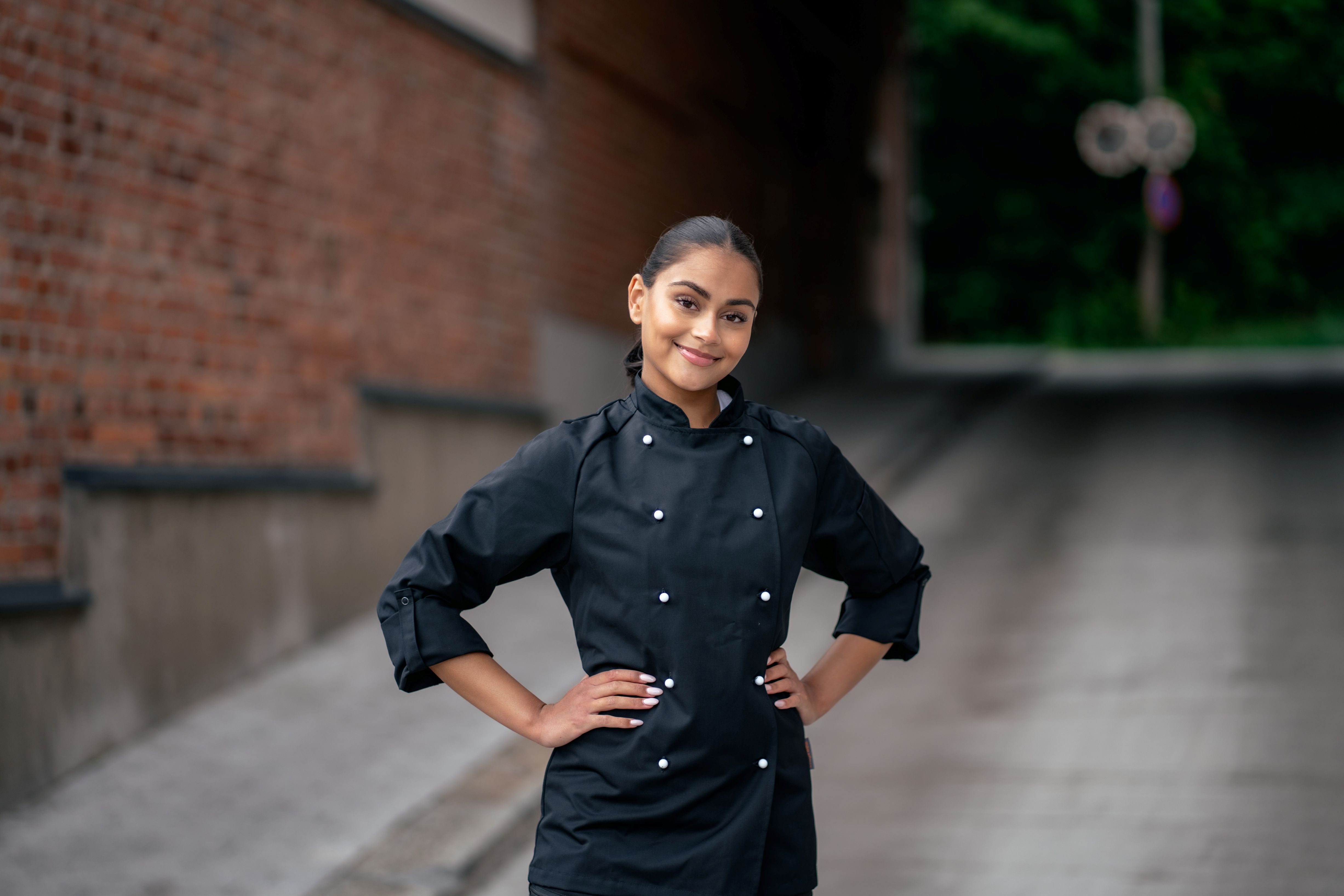 Kuvassa nainen seisoo ulkona kädet vyötäröllä. Päällä naisella on Image Wearin Aarre-malliston kokkitakki mustana valkoisilla irtonapeilla ja mustat reisitaskuhousut. 