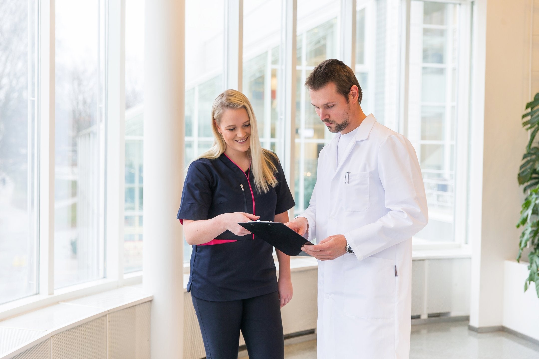 Kuvassa nainen seisoo aulassa päällään Image Wearin terveydenhuollon malliston valkoinen trikoopaita, turkoositummansininen liivi ja tummansiniset housut. 