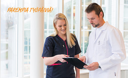 Kuvassa nainen seisoo aulassa päällään Image Wearin terveydenhuollon malliston valkoinen trikoopaita, turkoositummansininen liivi ja tummansiniset housut. 