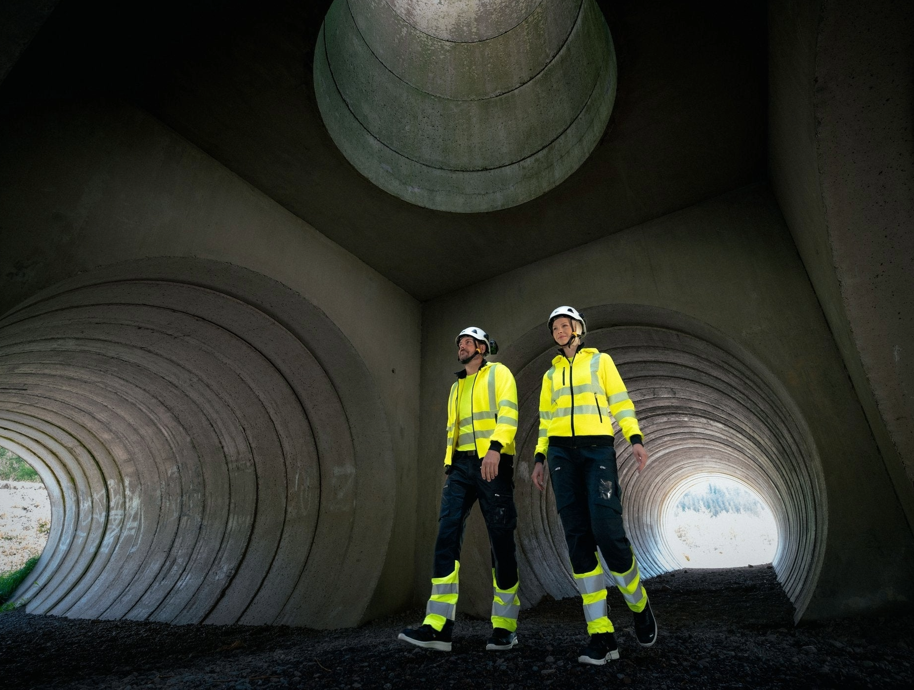 Kuvassa nainen ja mies kävelevät betonitunnelissa. Molemmilla on päällä Image Wearin kesämalliston keltaiset standardoidut varoitusvaatteet.