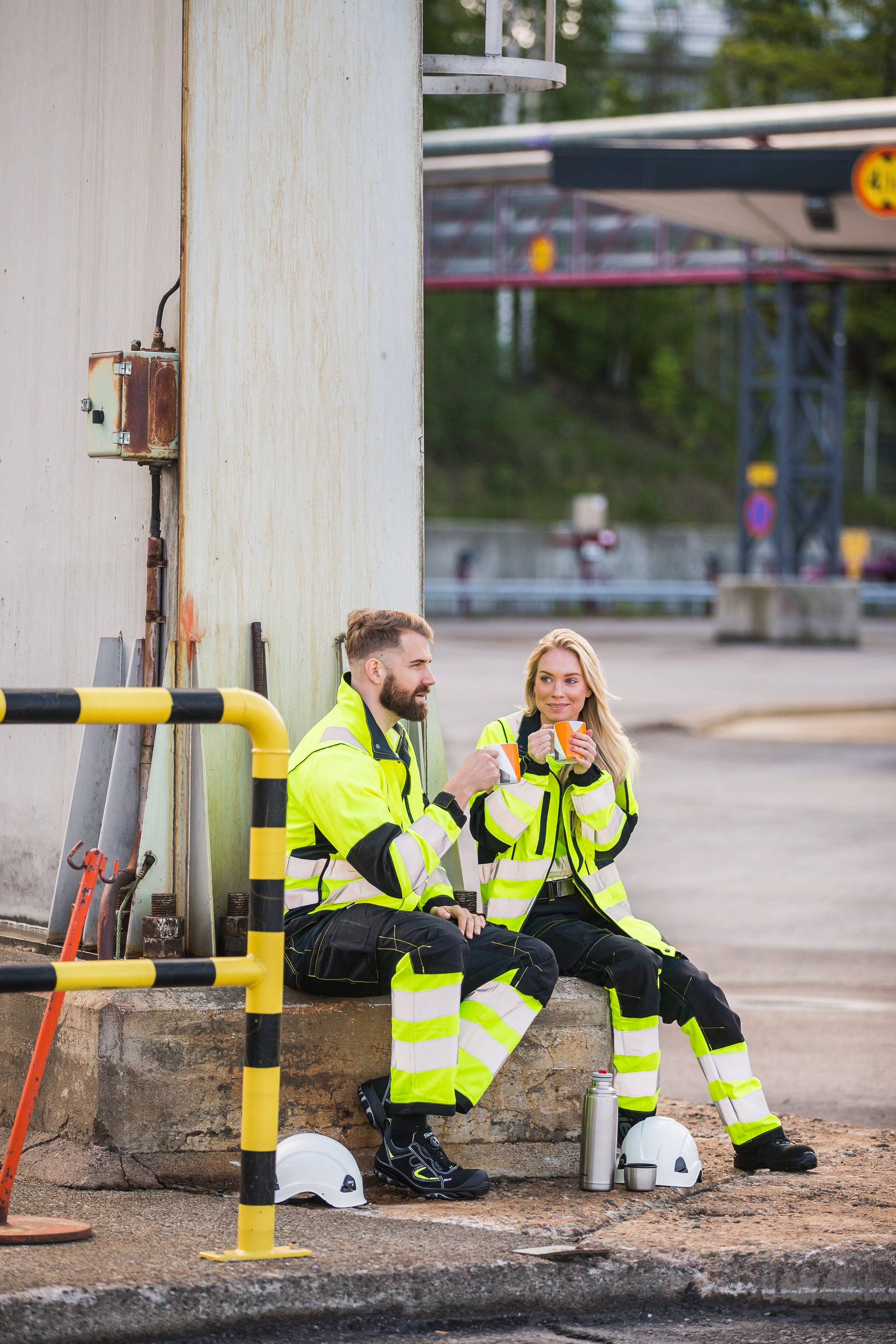 Kuvassa nainen ja mies istuvat ulkona kahvitauolla. Molemmilla on päällä Image Wearin huomioväriset keltaiset varoitusvaatteet ja jalassa Sievin turvakengät.