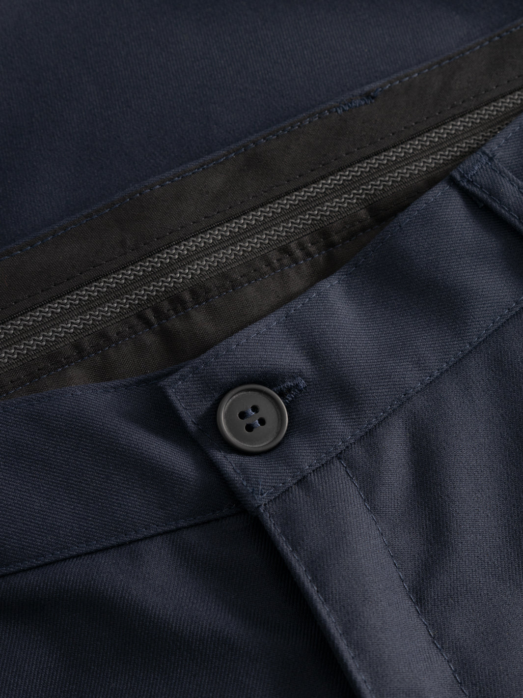 Yksityiskohtakuva tummansinisten housujen vyötärön nappikiinnityksestä ja vyötärökaitaleen sisäosasta. 