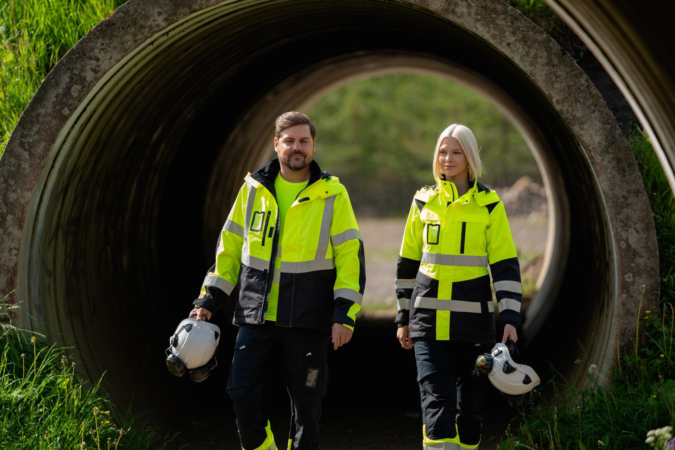 Kuvassa miehellä ja naisella ulkona päällä varoituskuoritakit, reisitaskuhousut ja varoituspaidat Image Wearin mallistosta sekä Petzlin valkoiset turvakypärät ja Sievin turvajalkineet.