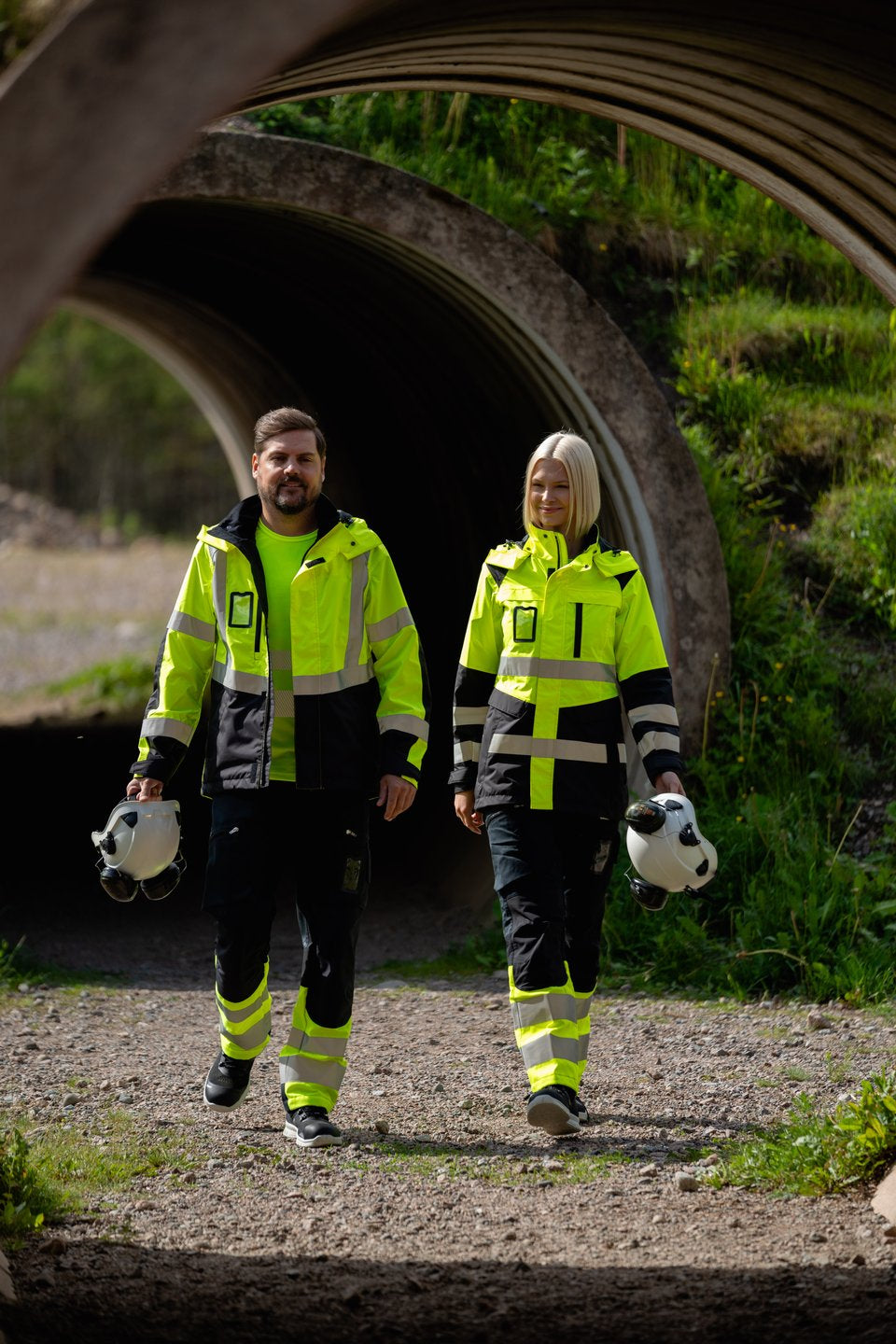 Kuvassa miehellä ja naisella ulkona päällä varoituskuoritakit, reisitaskuhousut ja varoituspaidat Image Wearin mallistosta sekä Petzlin valkoiset turvakypärät ja Sievin turvajalkineet.