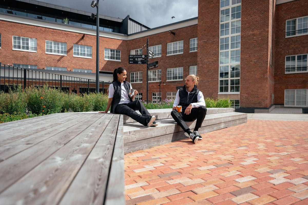 Kuvassa mies ja nainen istuvat ulkona. Molemmilla on päällään Image Wearin mustat reisitaskuhousut ja liivi sekä valkoinen t-paita. Jalassa Sievin turva-ja työkengät. 