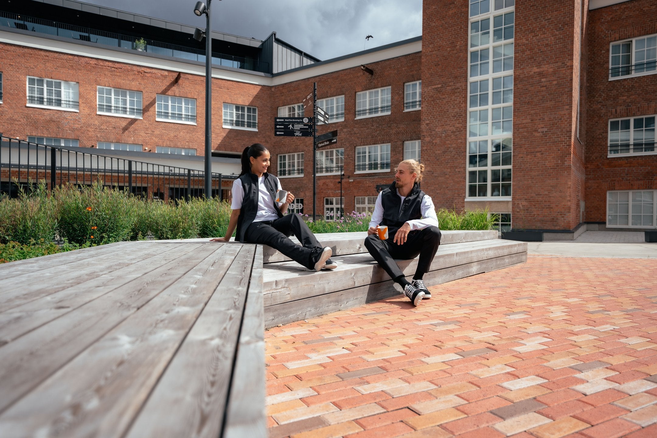 Kuvassa mies ja nainen istuvat ulkona. Molemmilla on päällään Image Wearin mustat reisitaskuhousut ja liivi sekä valkoinen t-paita. Jalassa Sievin turva-ja työkengät. 