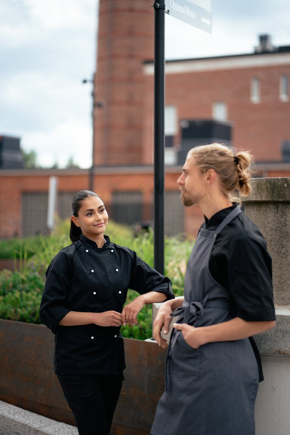 Kuvassa mies ja nainen seisovat ulkona. Molemmilla on päällään Image Wearin kokkitakit ja miehellä lisäksi siniharmaa suutarin esiliina. 